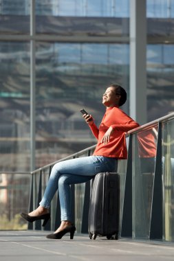Tam uzunlukta yan portresi mutlu genç Afro-Amerikan seyahat bavul cep telefonu ile istasyonunda oturan kadın