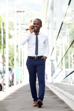 Tam vücut yürüme ve şehir üstünde hareket eden telefon ile sokak konuşurken gülümseyen Afrikalı-Amerikalı işadamı portresi 