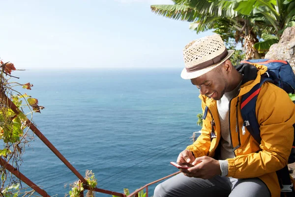 Portrait of happy african american travel man sitting by sea looking at mobile phone