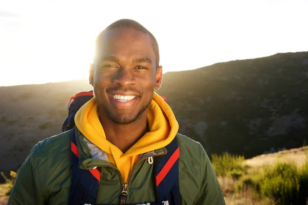 Close up portrait of handsome young african american man with back pack posing in nature with sunset in background