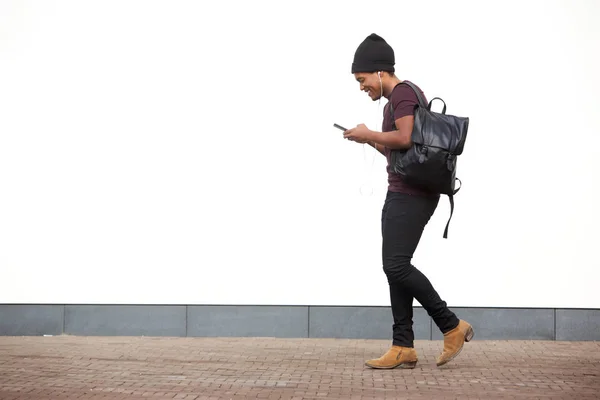 バッグを持って歩き スマートフォンで音楽を聴くアフリカ系アメリカ人男性の全身側面の肖像画 — ストック写真