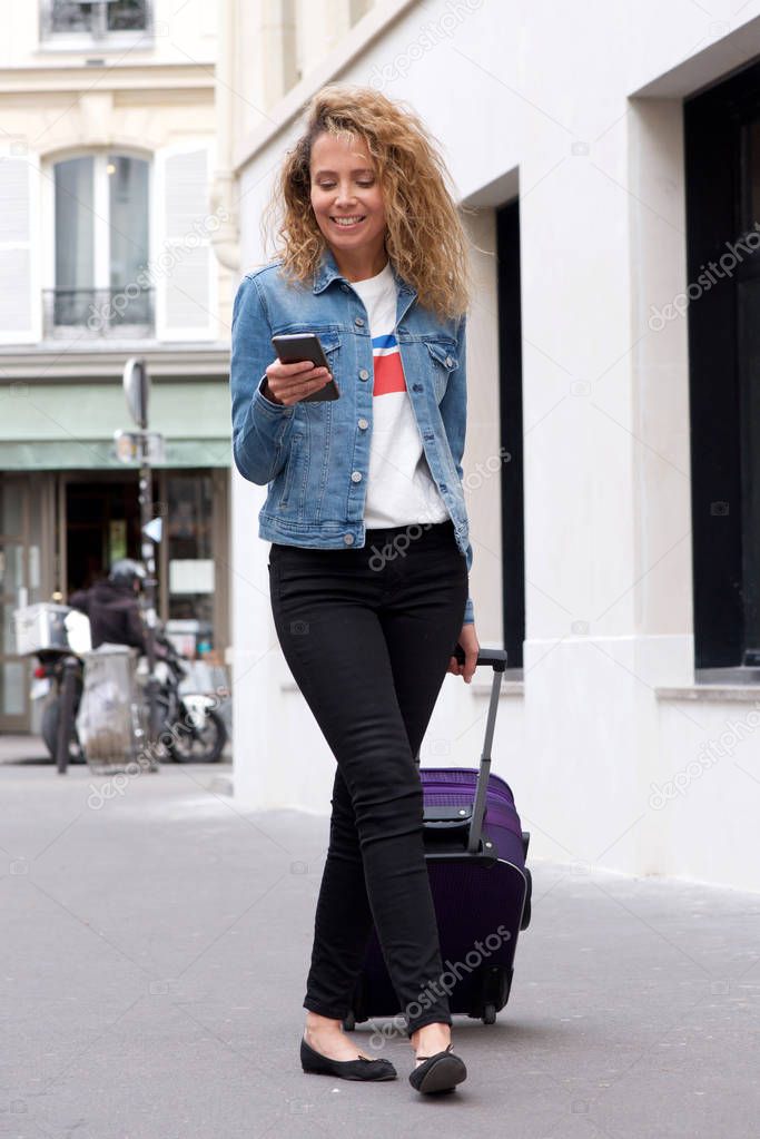 Full length portrait of happy middle age woman walking with mobile phone and suitcase on street