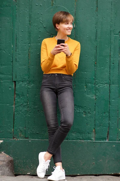 携帯電話で壁に寄りかかっている若い女性の全身肖像画 — ストック写真
