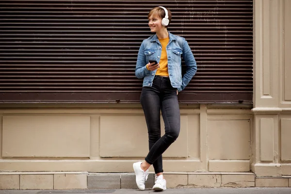 携帯電話とヘッドフォンで外に立つクールな若い女性のフルボディポートレート — ストック写真