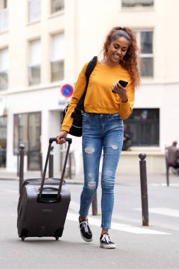 Şehirde bavul ve cep telefonu ile yürüyen mutlu genç kadının tam uzunlukta portre