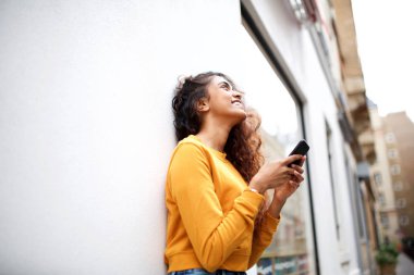 Beyaz duvara yaslanmış ve akıllı telefon tutan mutlu genç hintli kadının yan portresi