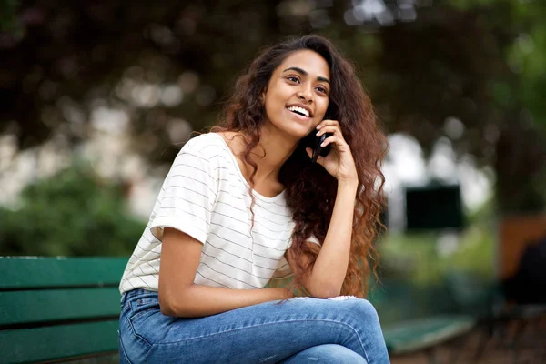 携帯電話で話す公園のベンチに座っている幸せな若いインドの女性のサイドポートレート — ストック写真