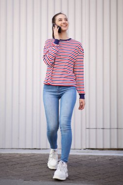 Gülümseyen genç kadının cep telefonuyla yürüyüp konuşmasının tam uzunlukta portresi