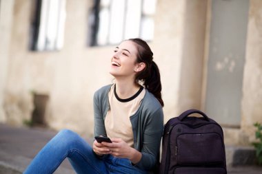 Çanta ve cep telefonu ile dışarıda oturan mutlu kız öğrenci portresi