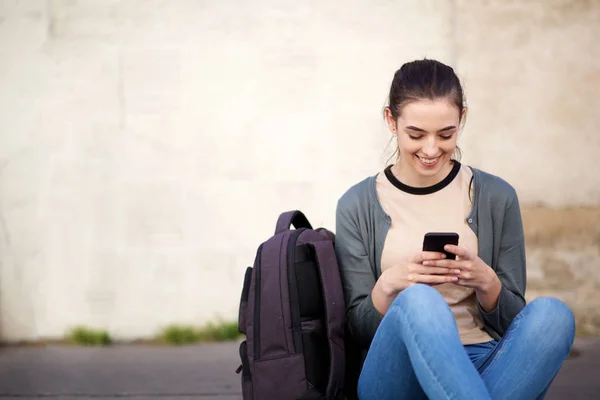 外の床に座って携帯電話を見ている笑顔の女子学生の肖像画 — ストック写真