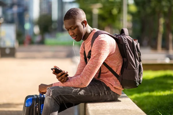 Portrait of young black travel man sitting outside with mobile phone and bags