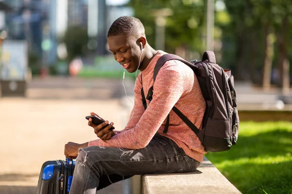 旅行バッグと携帯電話で外に座っている若いアフリカ人男性のサイドポートレート — ストック写真