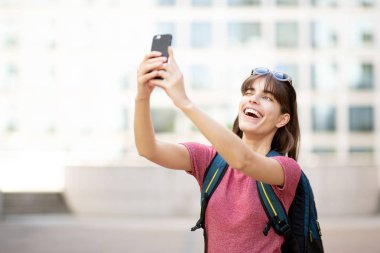Dışarıda cep telefonuyla selfie çeken mutlu genç bir kadının portresi.