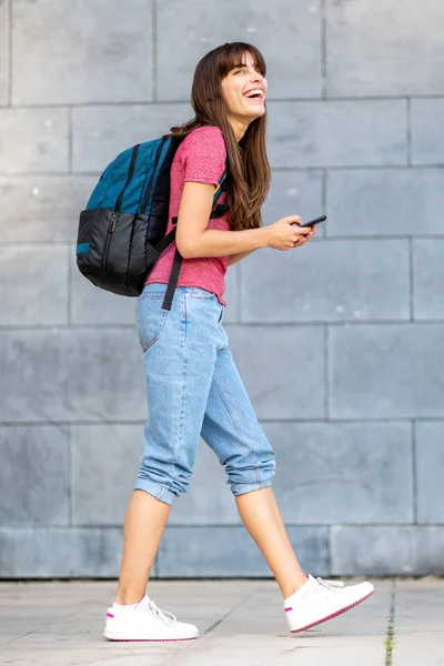 Full body profile portrait of young woman walking with cellphone and bag