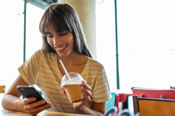 Портрет Улыбающейся Молодой Женщины Сидящей Кафе Пьющей Напиток Смотрящей Мобильный — стоковое фото
