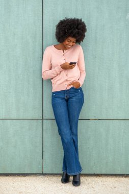 Yeşil duvara yaslanan ve cep telefonuna bakan mutlu, gülümseyen Afro-Amerikan kadının tüm vücut portresi.