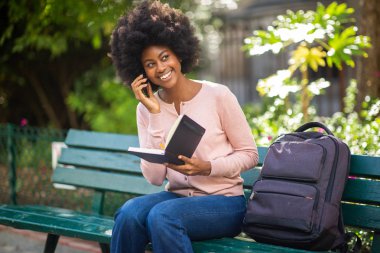 Mutlu gülümseyen zenci kadın portresi kitap çantasıyla dışarıda oturuyor ve telefonla konuşuyor.