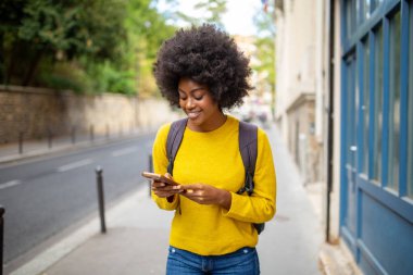 Şehirde cep telefonu ve çantasıyla dolaşan gülümseyen Afrikalı Amerikalı genç bir kadının portresi.