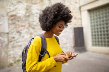 Şehirde çantalı ve cep telefonlu gülümseyen Afrikalı Amerikalı kadının yan portresi.