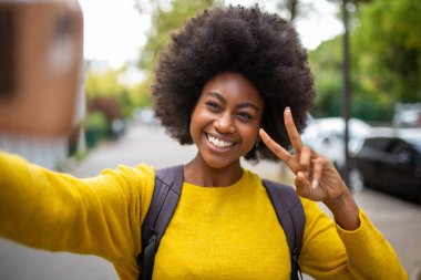 Dışarıda cep telefonuyla selfie çeken gülümseyen afro kadın portresi.