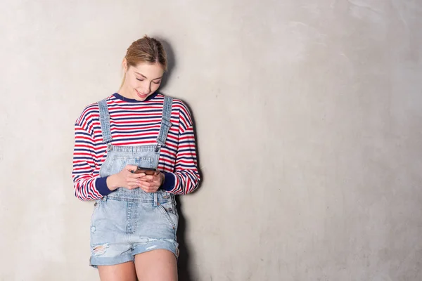 Portrait of blond girl smiling with cellphone by gray wall