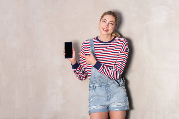 Retrato Una Chica Rubia Sonriente Sosteniendo Teléfono Celular Señalándolo —  Fotos de Stock