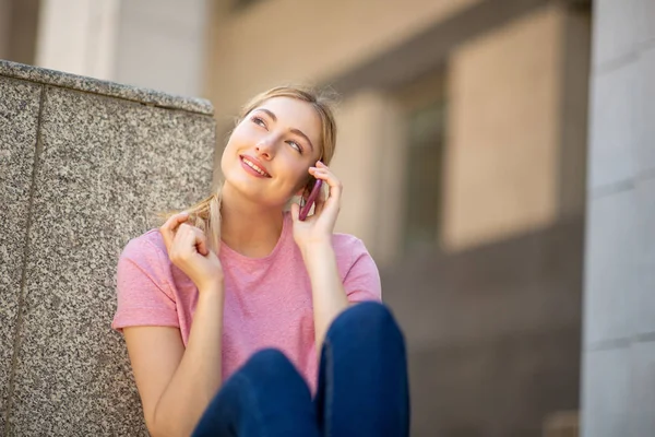 一个面带微笑的金发少女坐在外面用手机聊天的画像 — 图库照片