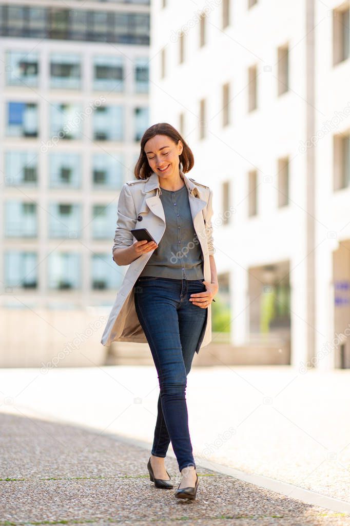 Full body portrait happy woman walking outside in city with cellphone