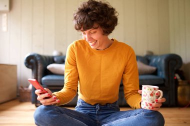 Evde oturup cep telefonuna bakan gülümseyen bir kadının portresi. 