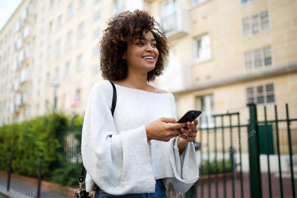 Portrait of smiling young african american woman walking in city with mobile phone in hand
