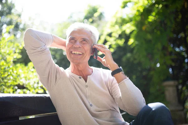 Portrait happy elderly man sitting on park bench talking with cellphone