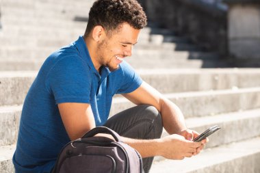 Genç melez ırkın portresi. Merdivenlerde çantasıyla oturmuş telefona bakıyor.