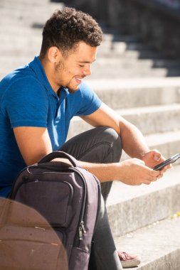 Merdivenlerde çantasıyla oturan ve cep telefonuna bakan Afro-Amerikan genç bir adamın yan portresi.