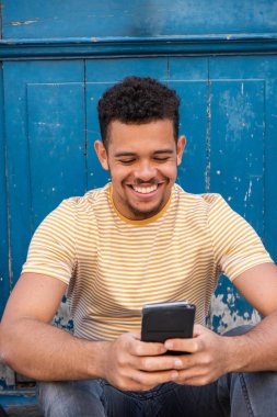 Mavi duvarda oturmuş cep telefonuna bakan gülümseyen melez genç bir adamın portresi. 