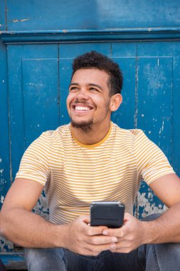 Mavi duvarın kenarında oturan, elinde cep telefonu tutan ve yüzünü çeviren mutlu, genç bir ırkın portresi.