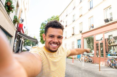 Portre gülümseyen genç melez adam şehrin sokaklarında selfie çekiyor ve işaret ediyor.