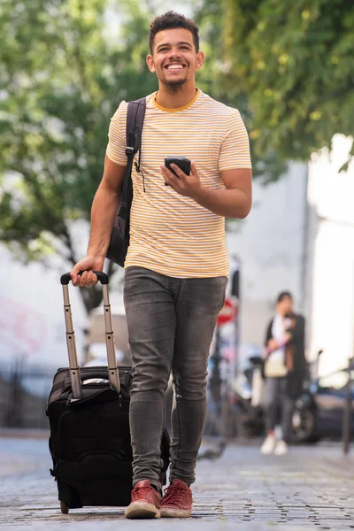Full body portrait of happy young man traveling with mobile phone and suitcase on street