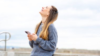 Mavi kazaklı genç bir kadın gökyüzüne bakıp elinde bir telefon tutarken ışıldıyor.