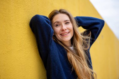 Gülümseyen kadın, elleri başının arkasında, sarı duvara yaslanmış.