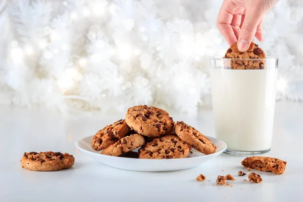 Tabakta el yapımı çikolatalı kurabiyeler. Bir kadının eli, Noel günü kurabiyeyi bir bardak taze süte batırır. Işıklar beyaz Noel ağacını süslüyor. Noel tatili kavramı.