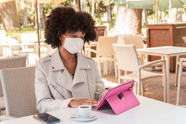 Young African American woman with curly hair and face mask works with her tablet while drinking coffee in an outdoor cafe. Working outside the office or telecommuting. College or high school student.