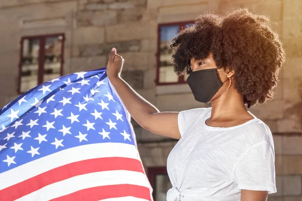 Maskeli ve afro saçlı siyahi kadın güneş ışığı altında Amerikan bayrağını tutuyor. Vatanseverlik ve ırkçılığa karşı mücadele.