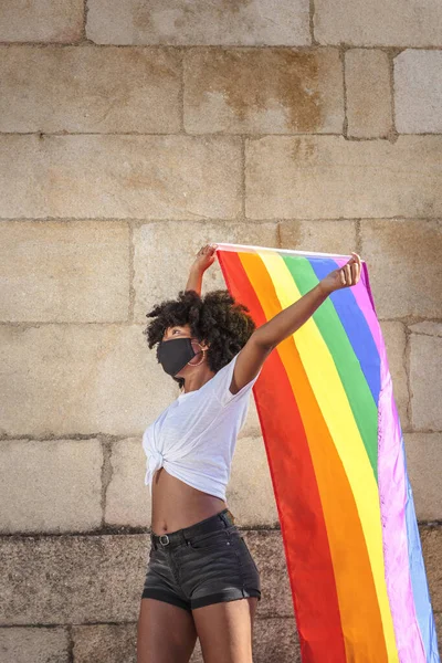 Yüz maskeli ve afro saçlı Afro-Amerikalı bir kadın, lgbtq bayrağını günışığının altında tutuyor. Cinsel özgürlük için savaşıyor..