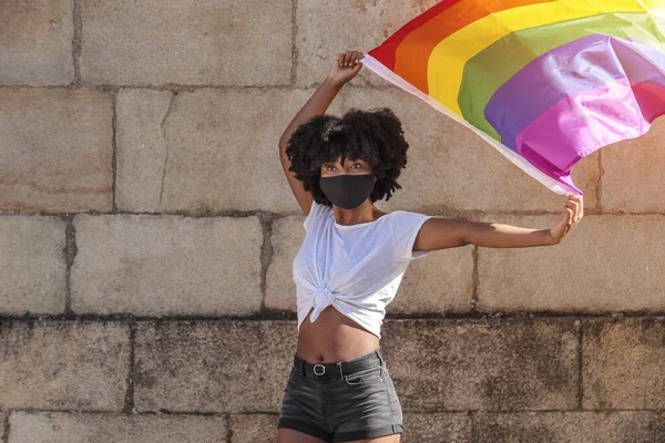 African American woman with a face mask and afro hair holds the lgbtq flag of gay pride under the sunlight. She fights for sexual freedom.