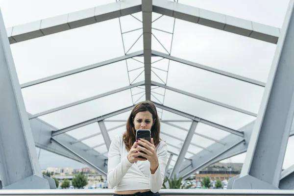 Frontal view of a young woman using the smartphone with a metallic structure in background