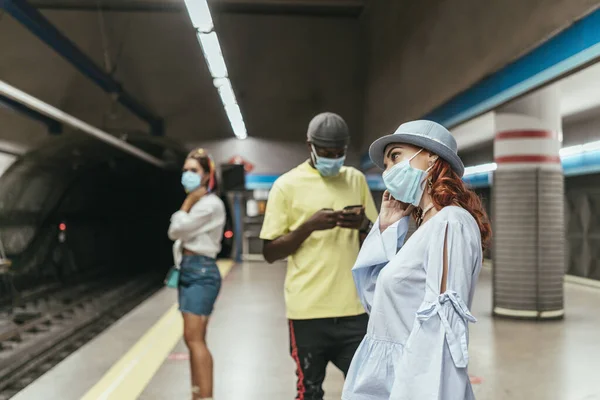 Interracial group of people with surgical mask waiting for the subway. There is black man between redhead and brunette women on underground station. Man is using smartphone.
