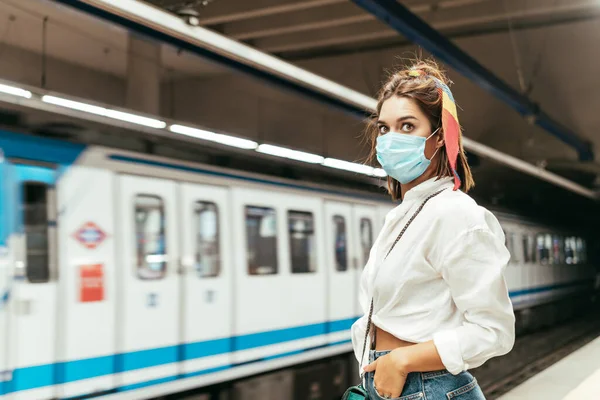 Woman looking to camera with blue surgical mask.She is wearing white shirt and jeans skirt. She is waiting in train station against underground train.