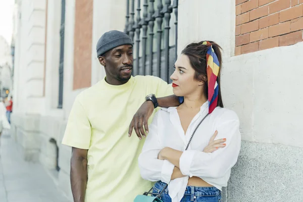 Black man and caucasian woman looking each other. She is with crossed arms and he is supported on girl. Wearing casual clothes. Wall on background