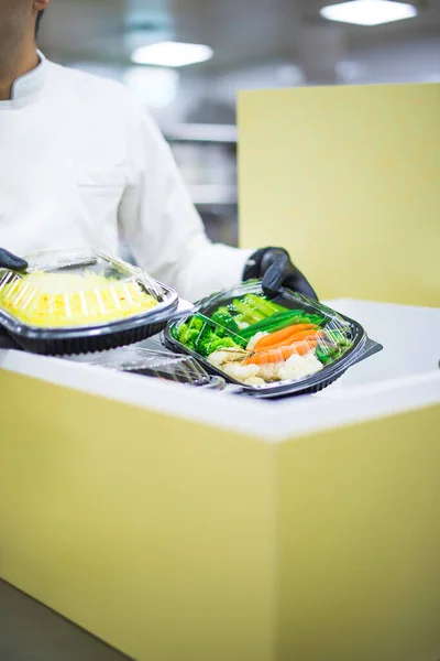 A close photo of the hands of a chef who puts fresh vegetables and cereals in a box. Food delivery in the boxes. Chef in white uniform and black gloves. Disposable plastic boxes.