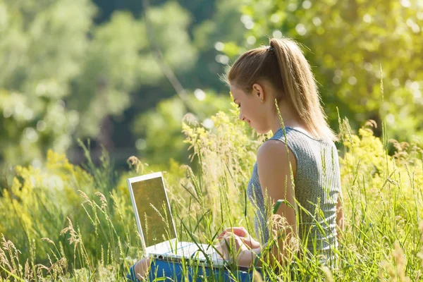 Ένα κορίτσι με ένα φορητό υπολογιστή στη φύση ανάμεσα σε καταπράσινο γρασίδι. — Φωτογραφία Αρχείου